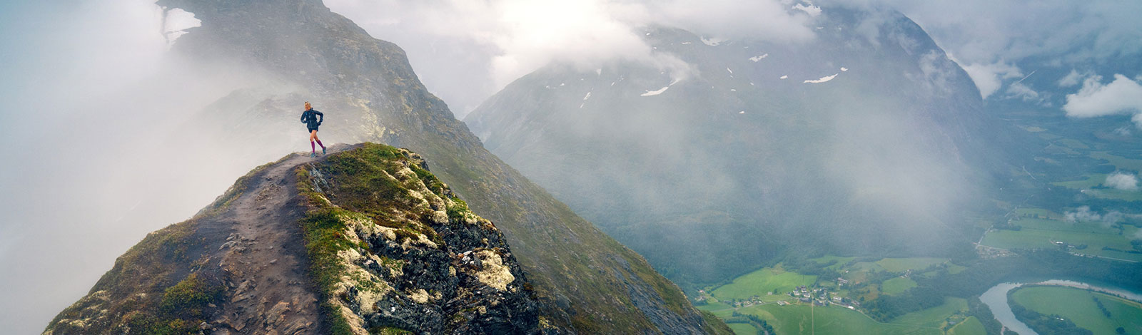 Skiny mountain landscape on the left means a woman runs over a narrow mountain path near the summit on the right, the valley can be seen with a small place and a river