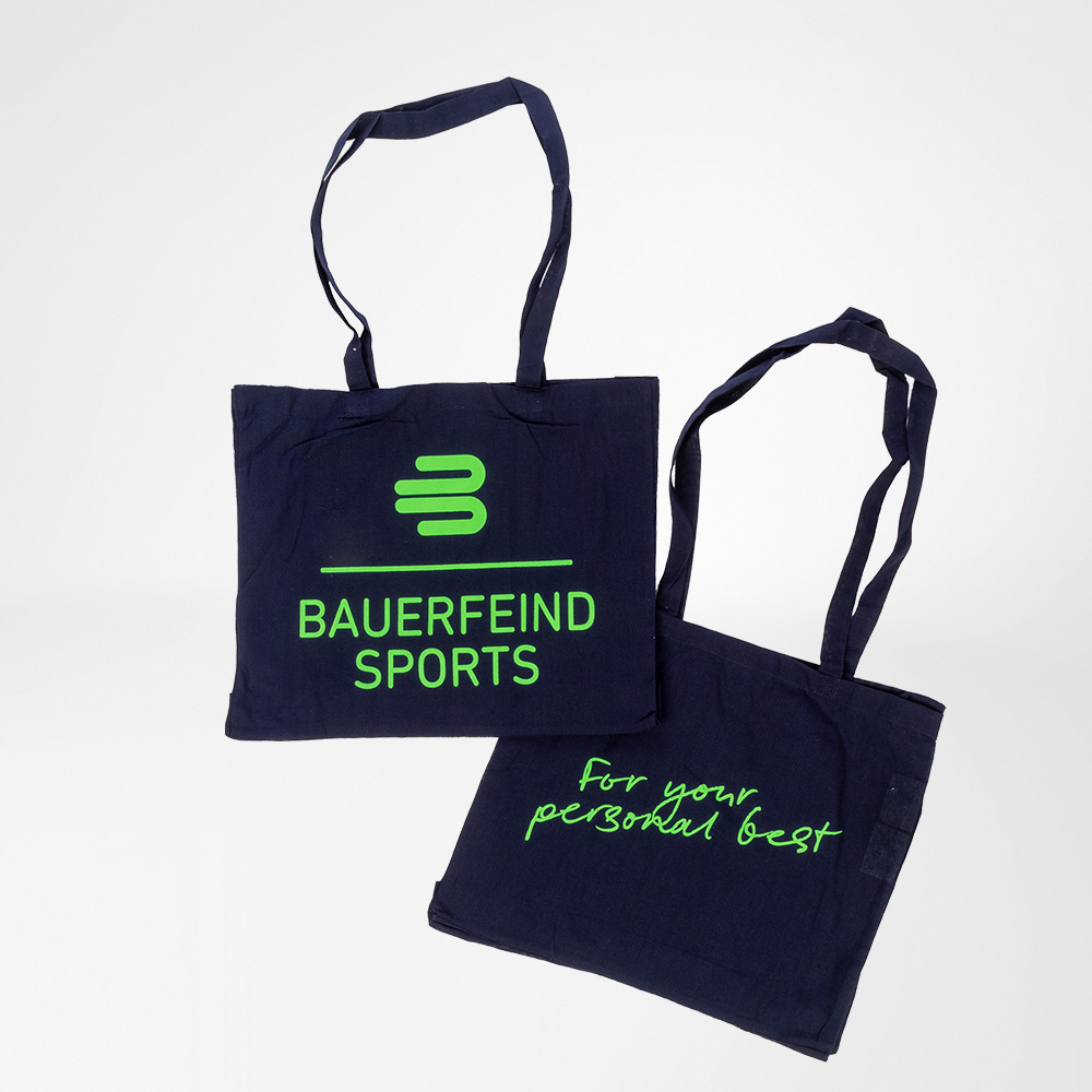 Two dark blue cotton tote bags by Bauerfeind Sports with bright green print. One displays the Bauerfeind Sports logo, the other the slogan "For your personal best". Both bags lie on a light surface.