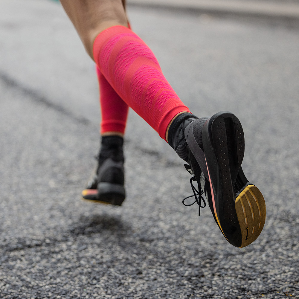 Close-up of a runner on asphalt, captured from behind in motion. She wears black running shoes and striking red Run Performance Sleeves with compression, providing targeted support for the calf muscles.