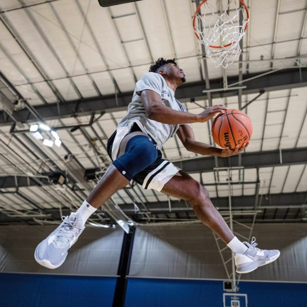 Flying high with full support – every jump matters! A basketball player in mid-air performing a dunk, wearing a blue knee brace on his right leg.
