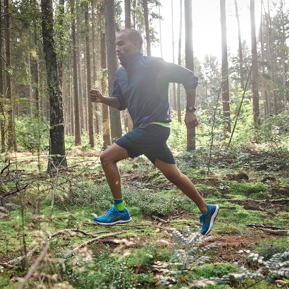 Run free – with stable support through uneven terrain. A man runs dynamically through a sunny forest. He wears a blue jacket, black shorts, and bright green calf sleeves.