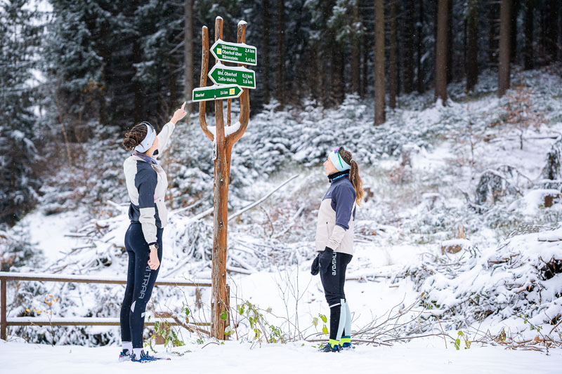 Zwei Frauen in Winterlaufkleidung stehen vor einem hölzernen Wegweiser im verschneiten Wald. Eine der Frauen zeigt mit dem Finger auf die Schilder, während die andere aufmerksam schaut. Der Wegweiser zeigt verschiedene Wanderrouten.