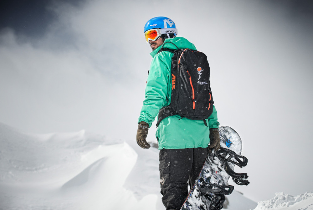 Snowboarders with a green jacket and board in hand looks over his shoulder backwards
