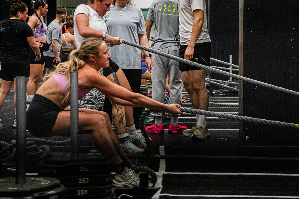 Athlete performing a sled pull during a HYROX competition, using a long rope to move a heavy sled across the indoor floor while staff and other participants stand near the workout stations in the background; the scene shows intense strength work