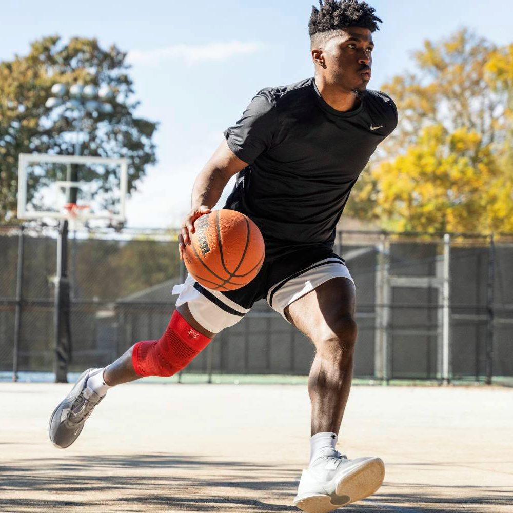 A basketball player wearing a red knee support sprints across the outdoor court while controlling the ball. The sunny day and trees in the background add a dynamic atmosphere to the scene.