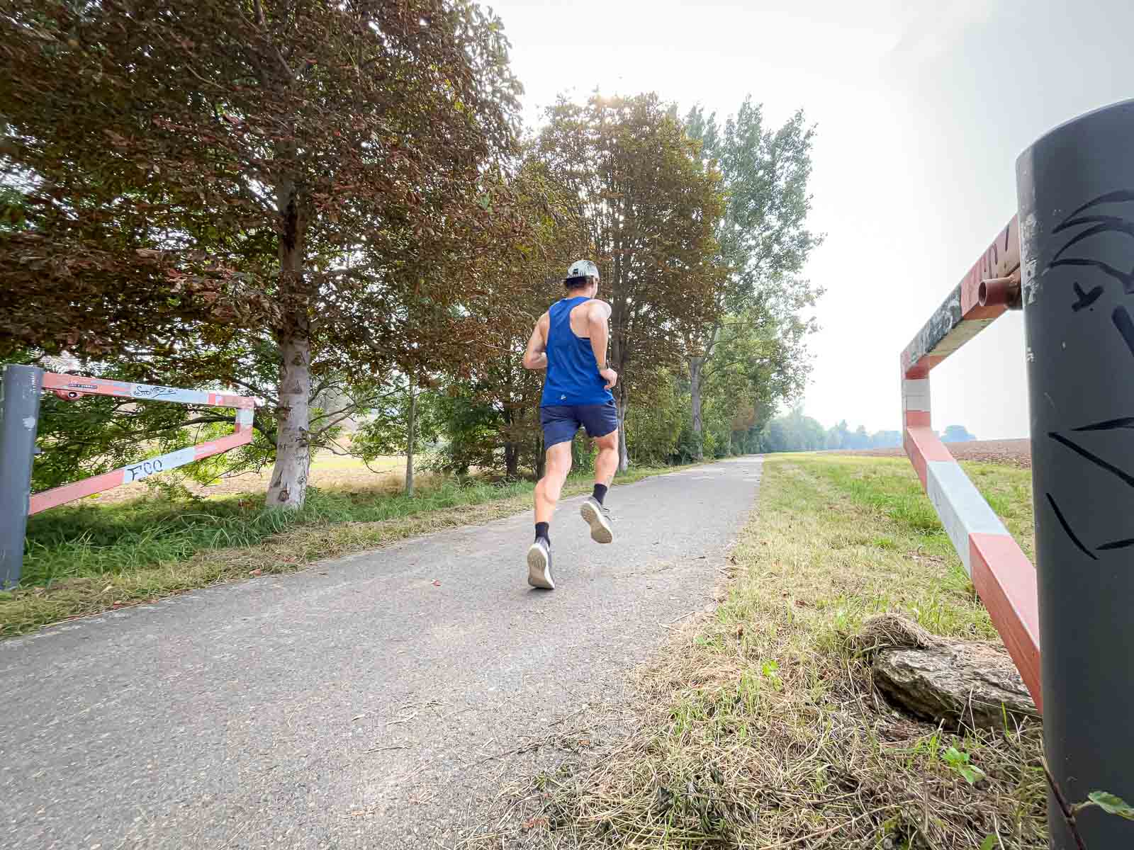 A runner in a blue singlet running on a paved path in nature. The path is lined with trees and a red-white barrier.