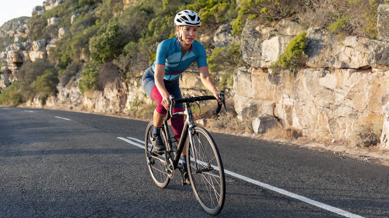 A woman with Knee Sleeves drives on a gravel path next to a meadow in the background a mountain rises on a gravel path