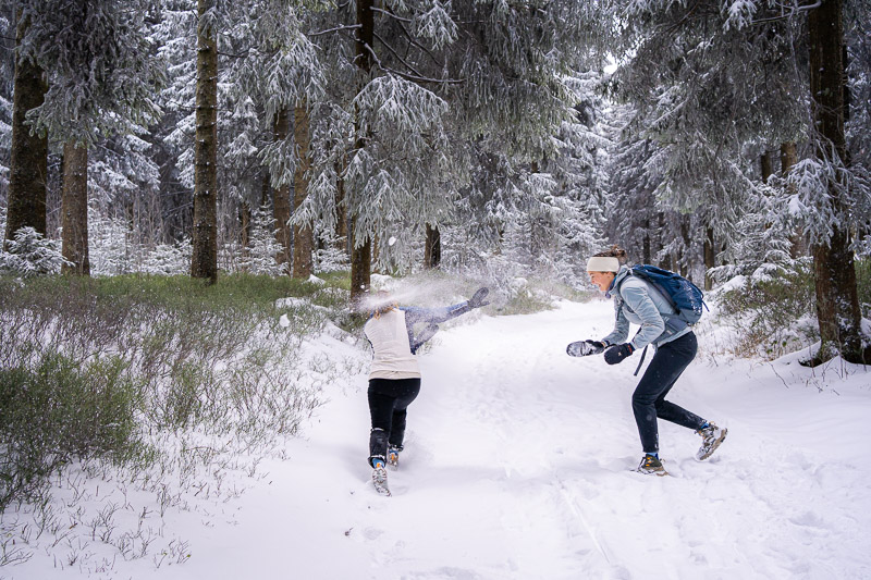 Zwei Frauen in warmer Winterkleidung haben eine Schneeballschlacht in einem tief verschneiten Wald. Die Frau rechts trägt eine graublaue Jacke und einen Rucksack, während sie gerade einen Schneeball wirft.
