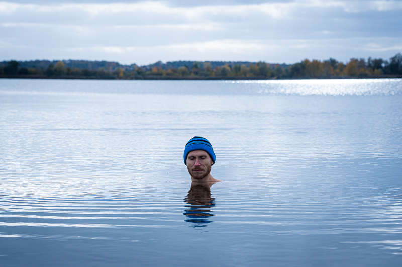 A man stands in a calm lake, submerged up to his neck, wearing a blue beanie, with the autumn landscape reflecting in the water.