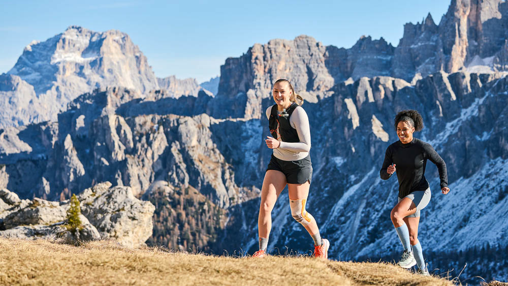 Two female runners train together on an open mountain ridge with a wide view of a rocky alpine landscape. One athlete wears an outdoor compression knee support. The image captures motion and outdoor performance.