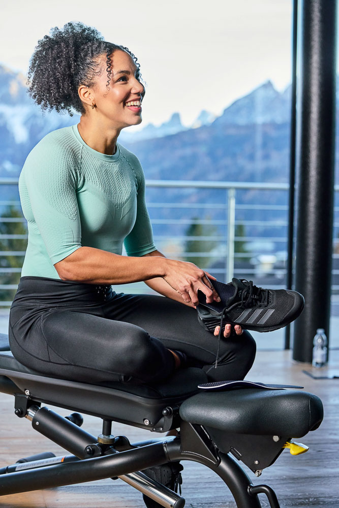 An athlete sits on a training bench tying a black sports shoe while smiling. She wears fitted sportswear, with large windows in the background overlooking an alpine landscape.