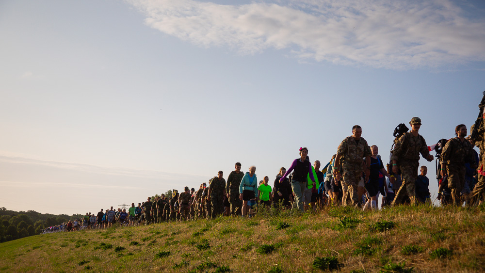 Hikers and soldiers in uniform walk in a long line across a dike during the 4Daagse in the morning light.