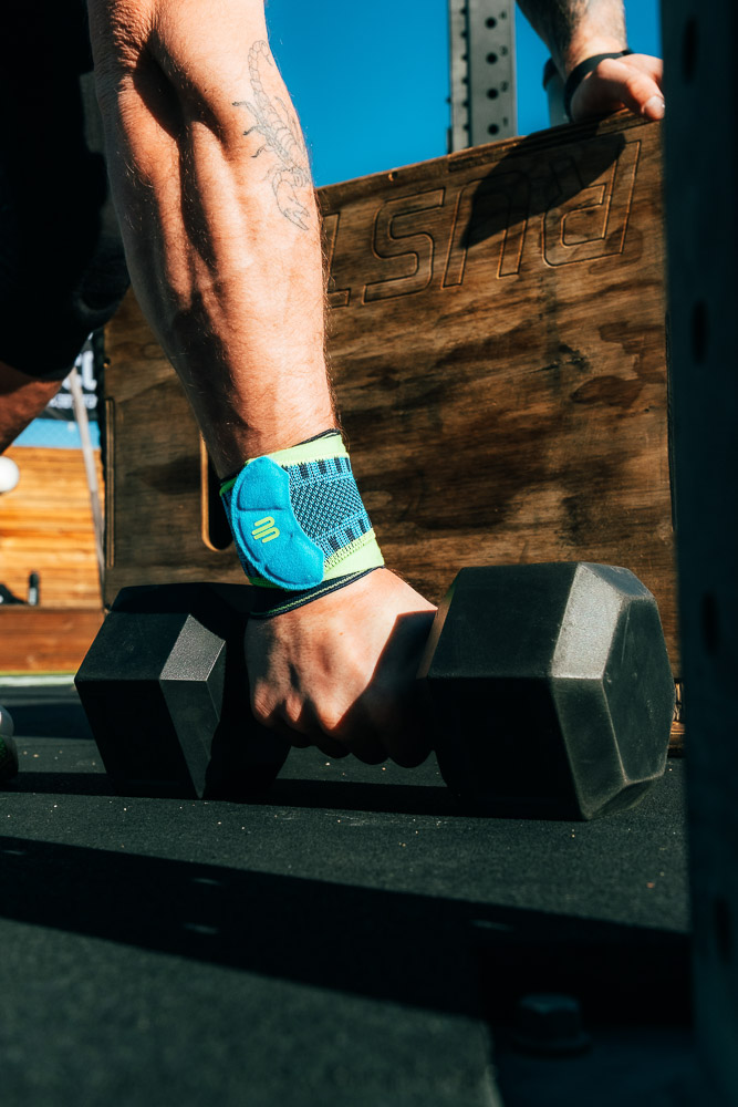 Close-up of a hand wearing a blue wrist support while gripping a black dumbbell; a wooden box and outdoor rack are visible in the sunlight in the background.