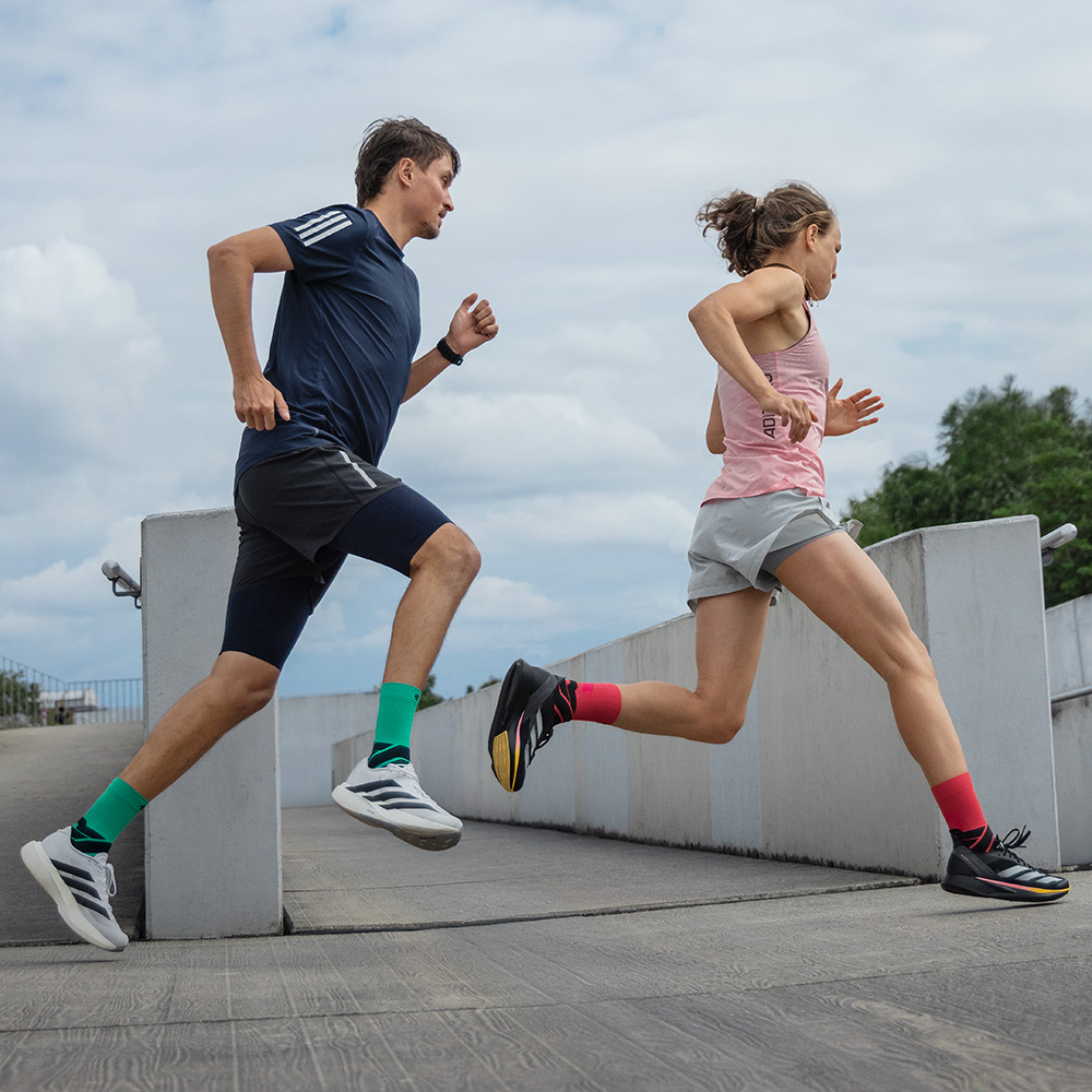 Two runners in motion on a ramp, a man in blue shorts with green Bauerfeind running socks and a woman in grey shorts with red Bauerfeind running socks.