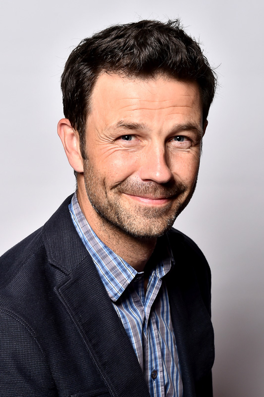 A man with short, dark hair and a warm smile wears a checkered shirt and a dark blazer. He looks directly at the camera against a neutral background.