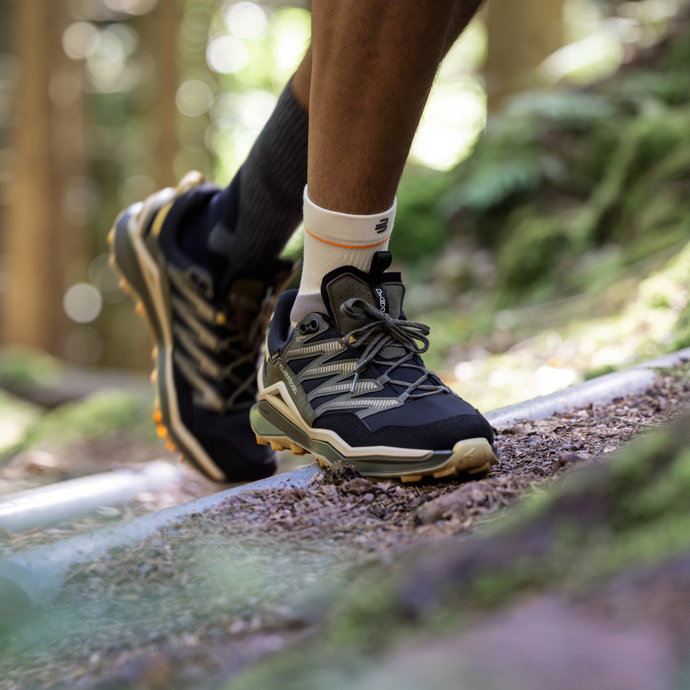 Close-up of a hiking boot on a root-covered forest trail. The Outdoor Compression Ankle Support is worn above the shoe, providing targeted compression around the ankle and support during foot roll-off on uneven surfaces.