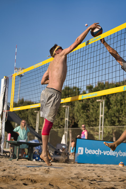 A beach volleyball player jumps at the net to block, while an opponent spikes the ball from the other side. The athlete is wearing a bright red compression sleeve on his right leg.