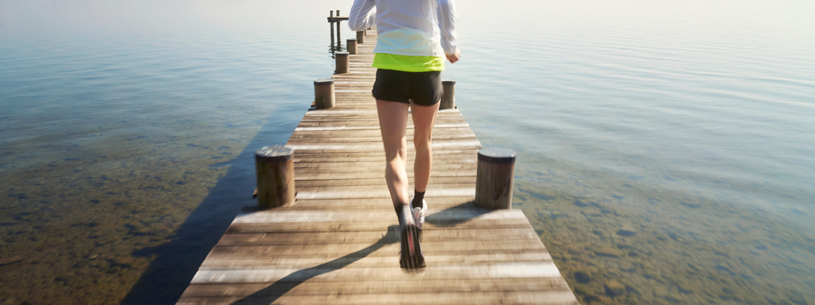 Woman with a white running jacket and short black running trousers runs over a footbridge towards the water