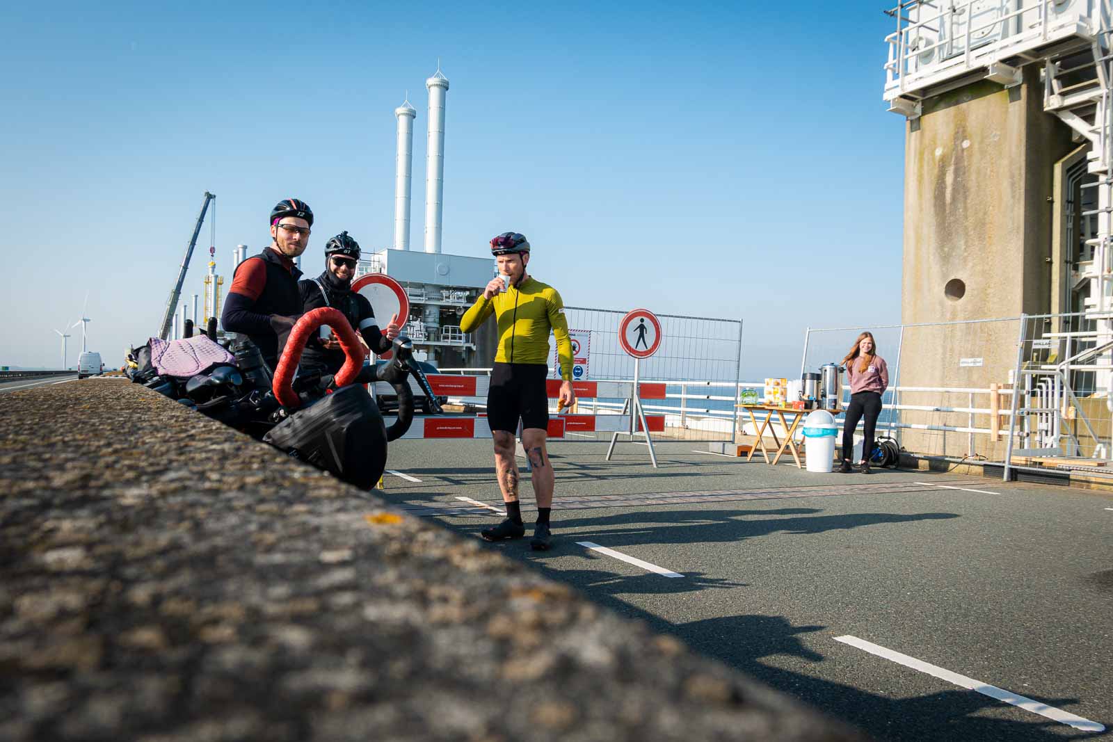 Three cyclists are waiting at a bridge construction site, drinking coffee and eating cookies