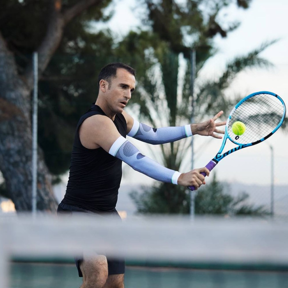 A tennis player wearing white compression sleeves hitting a forehand. Trees and a tennis court are visible in the background.