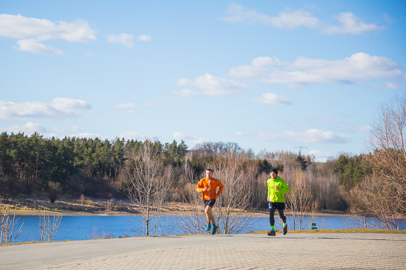 Two athletes running up a sunny hill with a view of a calm lake and a forested horizon. Both are wearing bright running jackets in orange and yellow.