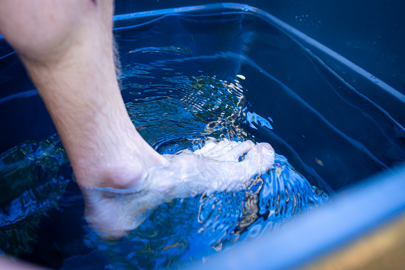 A foot dips into the cold water of a blue container. The clear water shows gentle ripples and light reflections on the surface.