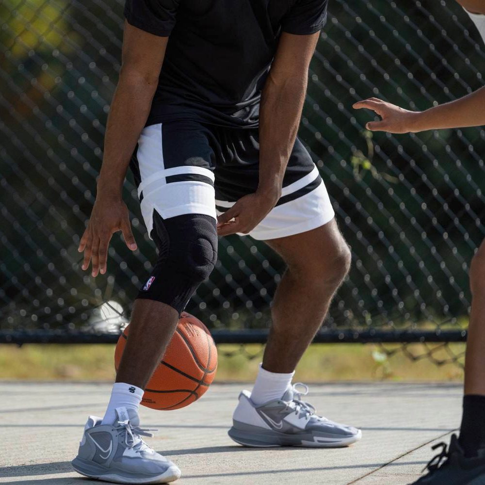 Close-up of a basketball player dribbling on an outdoor court. His right knee is supported by a black brace featuring an NBA logo. An opponent is partly visible in the frame.