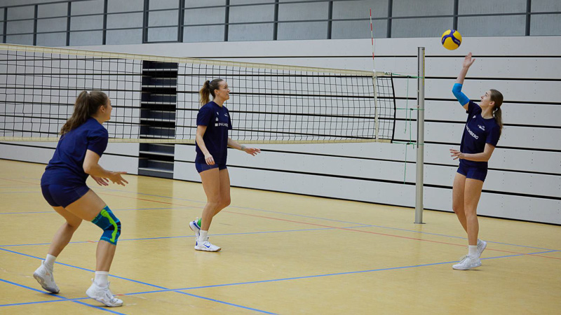 Three volleyball players on an indoor court during a game. One player wears a blue-green knee brace, another wears a blue arm support.