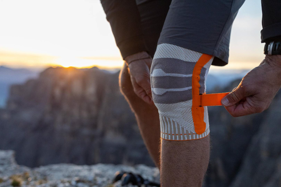 A hiker adjusts their orange knee brace during a mountain hike. Rocky mountains and a sunset are visible in the background.