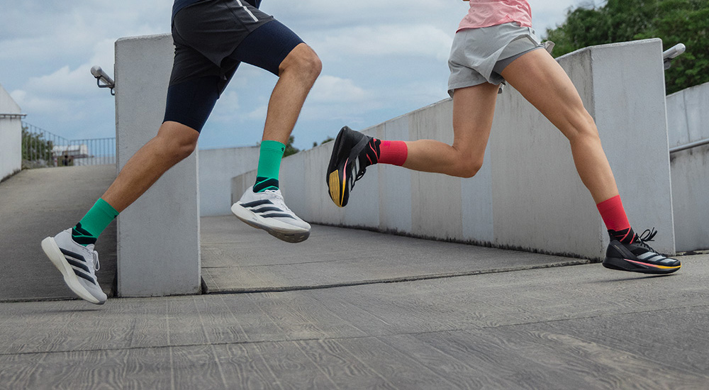 Two runners in motion sprinting up a ramp. The man wears green compression socks with white running shoes, while the woman sports black shoes and red compression socks.