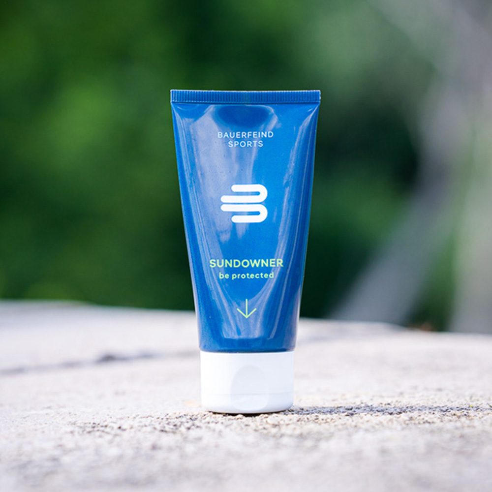 A blue tube of Bauerfeind Sports Sundowner sunscreen with SPF 30 is placed on a stone, surrounded by a green natural backdrop. The sunscreen provides water-resistant, vegan sun protection for outdoor activities.