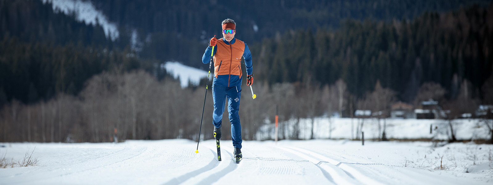 A man, photographed head-on from a distance, is cross-country skiing. Green forest trees can be seen in the background