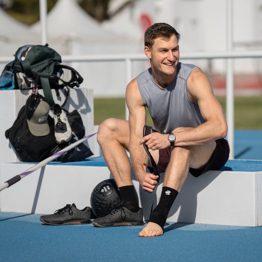 An athlete sits on a podium at an athletics facility, pulling a Bauerfeind Sports Ankle Support brace over his ankle. His sports shoes and a training ball are beside him.