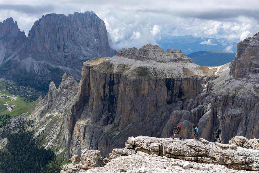 Landscape shot of many mountains in the Dolomites, with three hikers walking on the foreground peak.