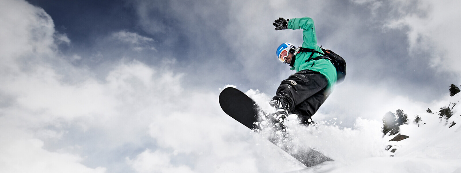 Frog perspective: Snowboarders in a green jacket comes from above over a crested board and much whispered snow can be seen