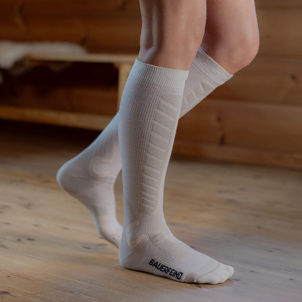 Perfect fit: White compression ski socks for maximum comfort on the slopes Close-up of white compression ski socks that reach just below the knees. The person stands on a wooden floor in a cabin. The socks offer extra comfort and support with their padded zones for skiing.