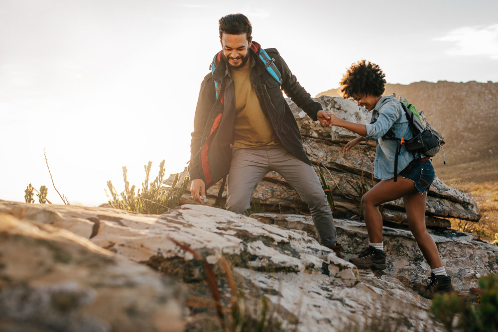 A young couple climbs a rocky slope together during sunset. The man helps the woman by reaching out his hand. Both wear backpacks and weatherproof hiking gear.