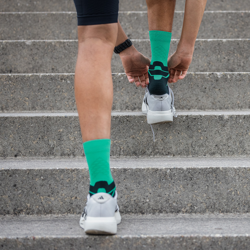 Rear view of a runner walking up stairs wearing green Bauerfeind running socks inside sports shoes. The focus is on the heel and the anatomical design of the sock.