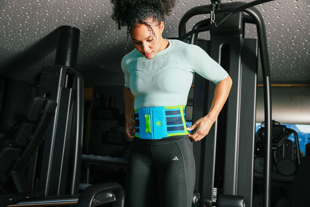 An athlete stands in a gym adjusting a blue back support around her waist, wearing functional sportswear and preparing for strength training.