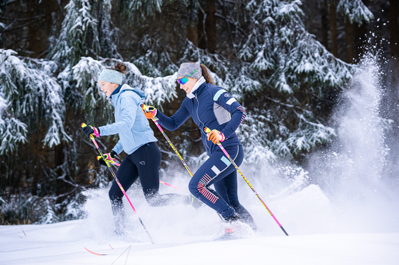 Two women cross-country skiing through snow-covered forests. They are wearing colorful winter sportswear and gliding dynamically through the snow. The scene is dominated by dense snowy fir trees, emphasizing the winter wonderland.