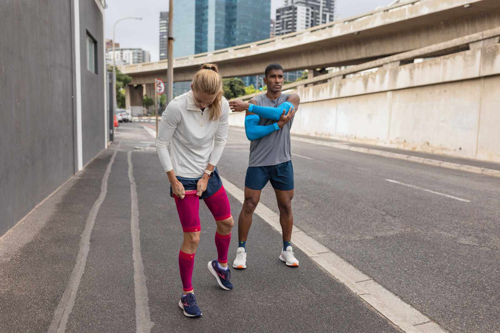 Also look good for the break: No problem with our stylish products A woman with pink compression Sleeves and a man with blue arm sleeves and short running socks take a break on the roadside of a big city and stretch out