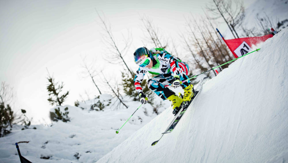 Ski runner in a blue and white-black racing suit drives down in the background a steep shaft in the background a red gate can be seen