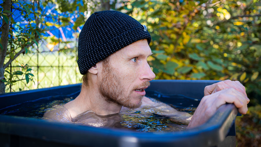 A man sitting in a black water container, surrounded by an autumn garden landscape. He is wearing a black knitted hat and looking intently to the side. The water reaches up to his chest, and the container is placed outdoors.