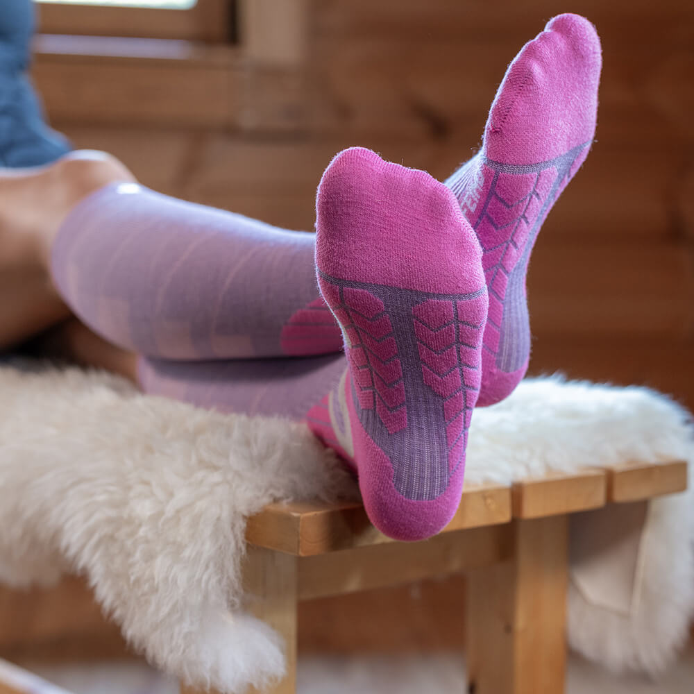 A person relaxes on a bench, wearing lilac compression ski socks with pink soles. The feet are stretched forward, showing the padded sole pattern. The scene takes place in a cozy cabin.
