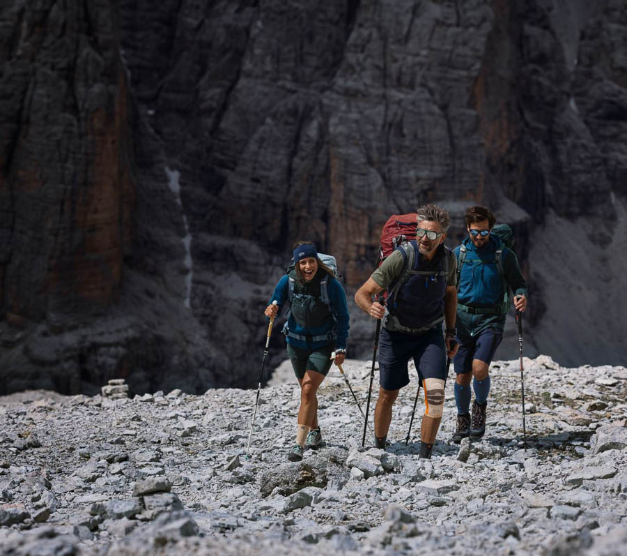 A group of three hikers, equipped with backpacks and trekking poles, climbing over rocky terrain in the mountains. One of the hikers is wearing a knee brace. Steep rock faces are visible in the background.