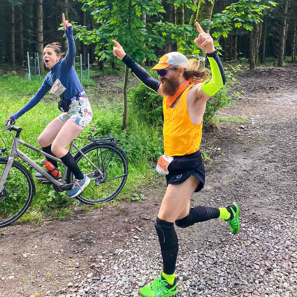 A man runs cheeringly to the finish line while a woman rides cheeringly alongside on her bike
