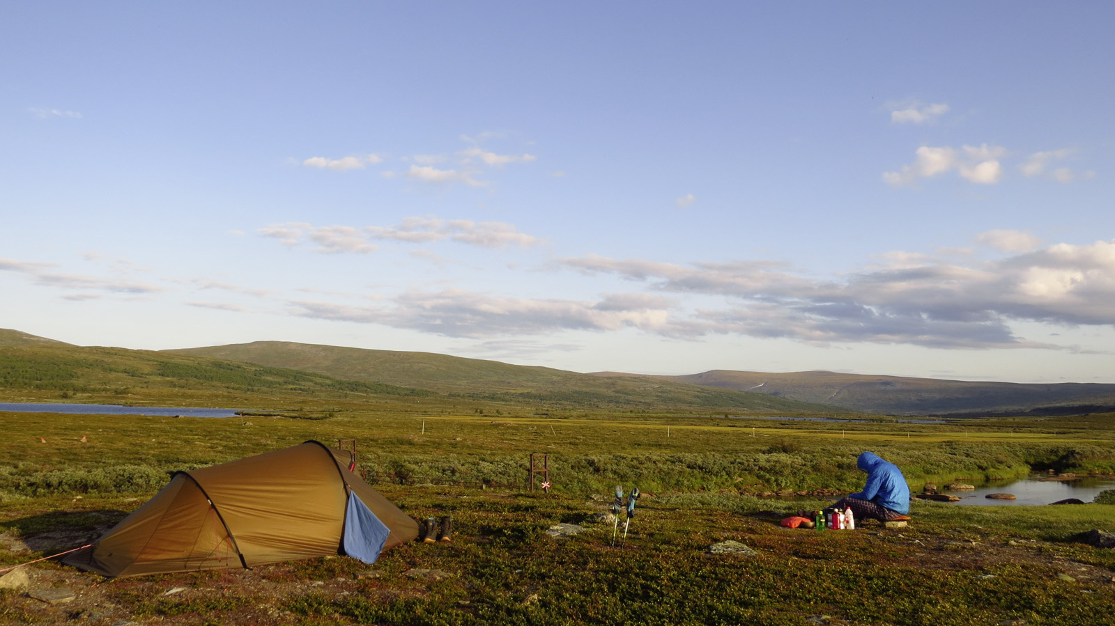 Man with a hooded jacket sits in front of a tent in a landscape in front of a blue sky