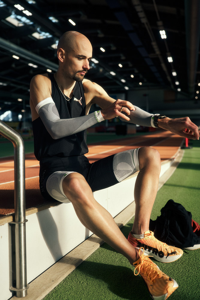 Marathon runner Hendrik Pfeiffer sits at the edge of an indoor track, putting on compression arm sleeves as part of his race preparation.