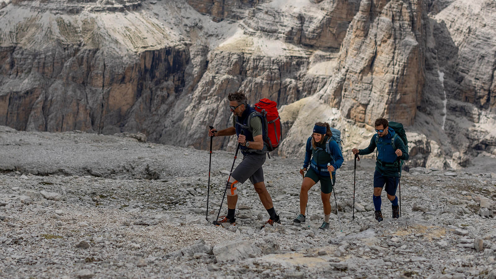 A group of three hikers in the Dolomites walking across a rocky field in a mountain panorama.