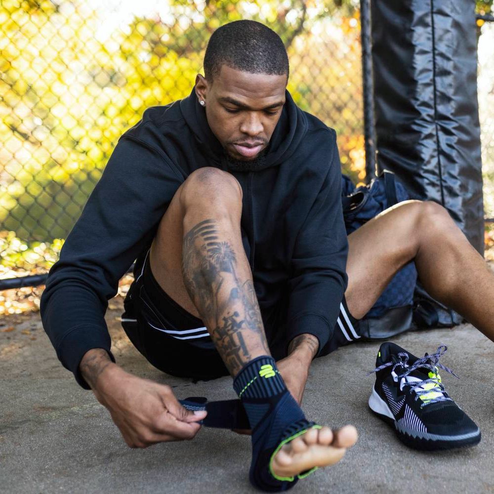Support where it counts – getting ready for action! A sportsman sits outdoors, wrapping a Bauerfeind Sports Ankle Support brace around his ankle. His sports shoes are nearby, with a metal fence in the background.